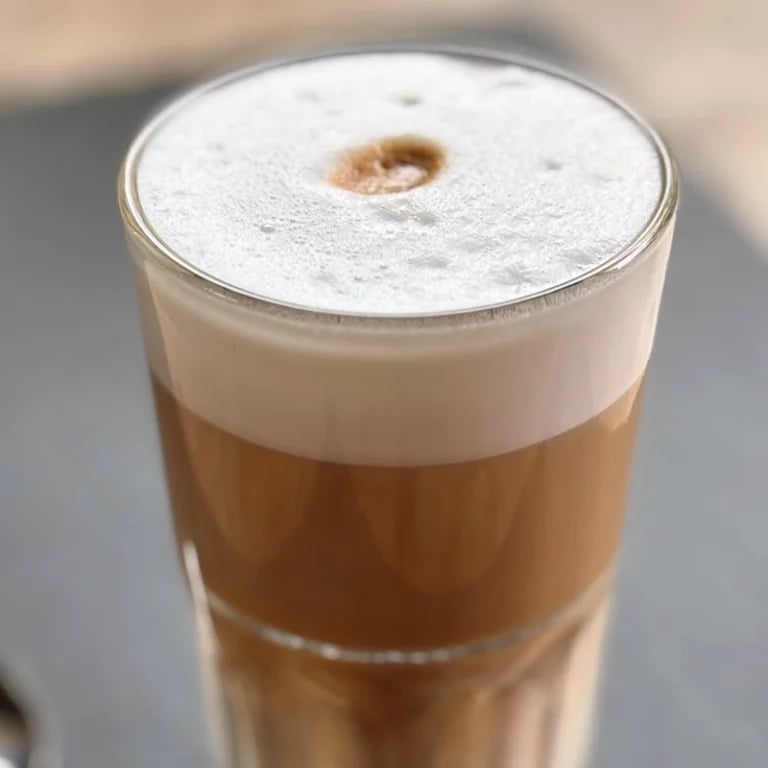 Tall glass of layered coffee drink with thick white foam top and small hole in center