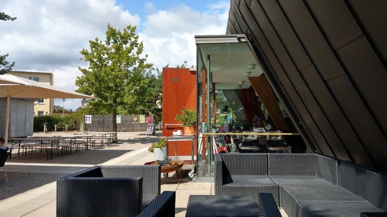 Modern outdoor patio area with contemporary architecture, seating, orange accent building, and landscaping under partly cloudy sky