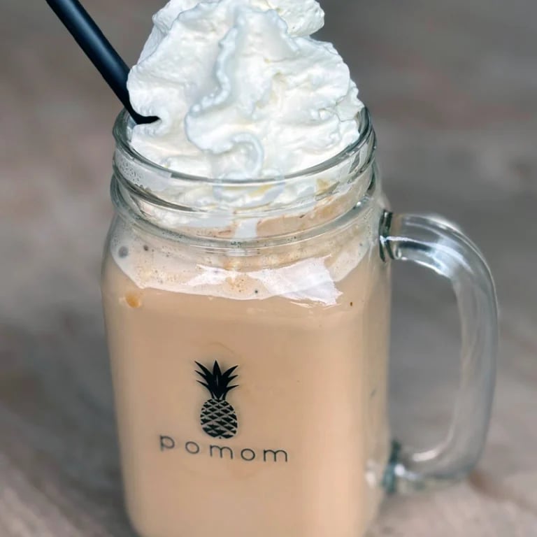 Iced beverage in a glass jar with whipped cream topping and a black straw, featuring a pomom pineapple logo