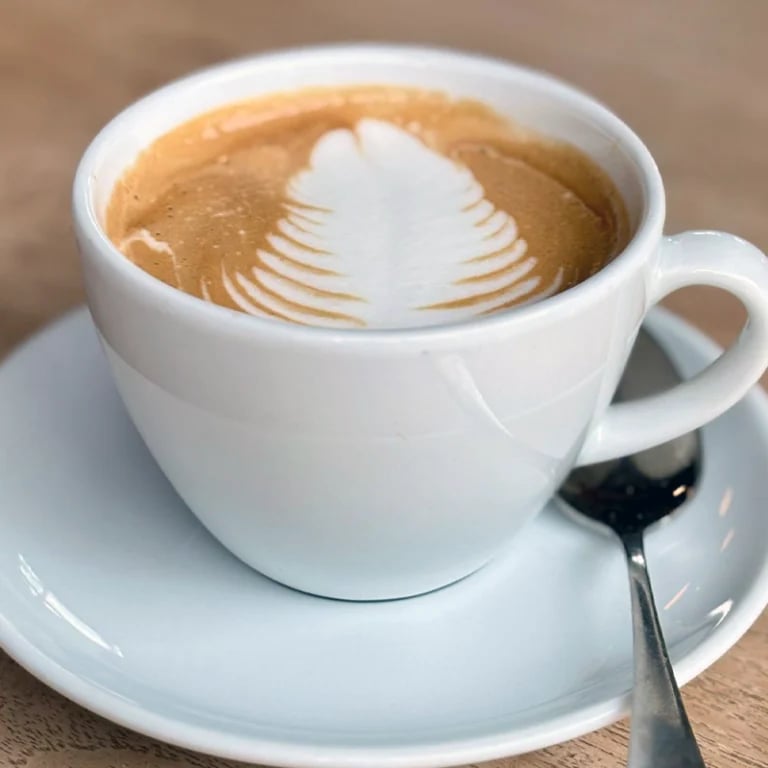 White cup of latte with decorative leaf latte art on saucer with spoon on wooden surface