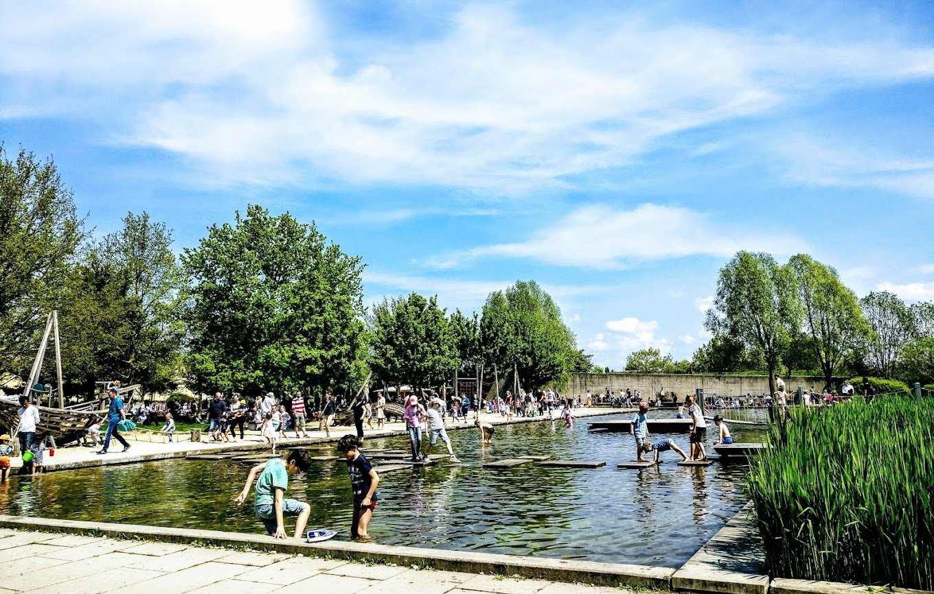 Crowds of people enjoying a sunny day at a waterfront park with trees, a canal, and a sailboat visible in the background