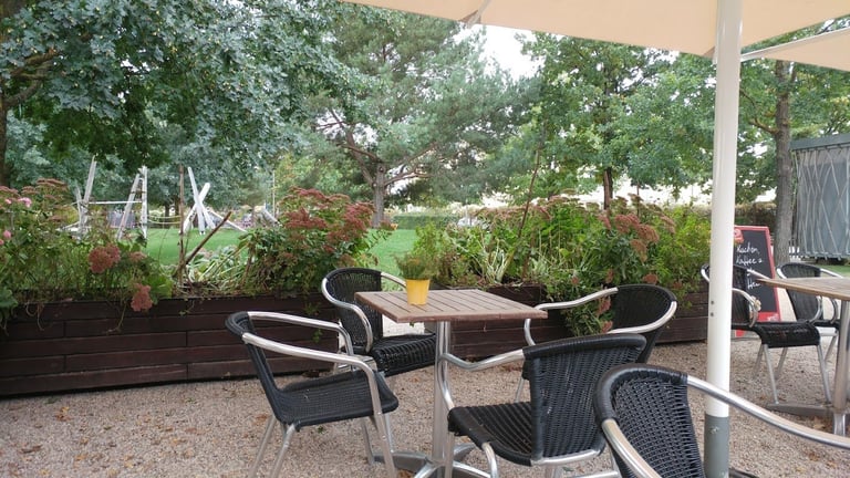 Outdoor patio area with black wicker chairs around a wooden table under a tan shade structure, surrounded by potted plants and lush greenery