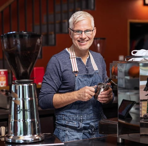 Smiling man with white hair and glasses wearing a denim apron, holding coffee grounds in front of brewing equipment