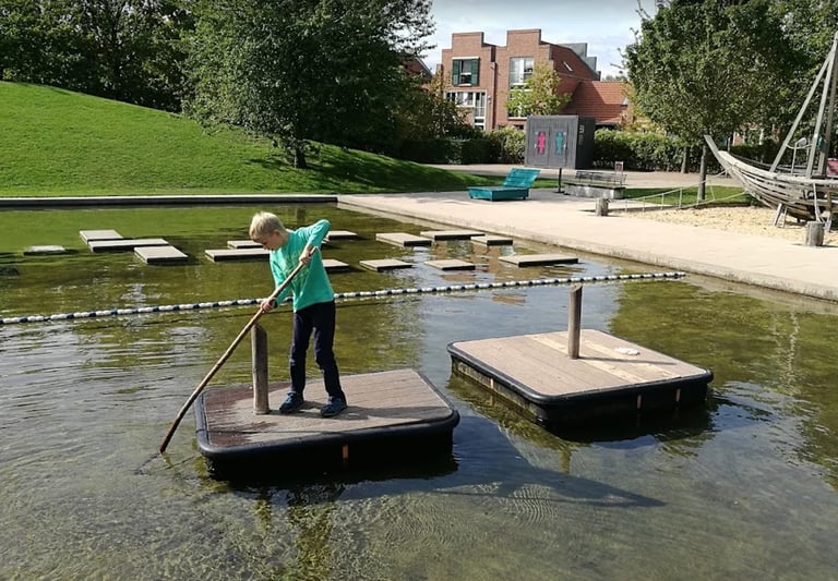 Child in green jacket standing on floating platform using pole vault over water in park