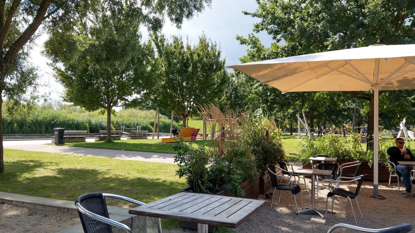Outdoor park seating area with tables, chairs, and beige umbrella shading a patio beneath green trees on a sunny day