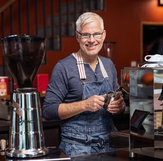 Smiling man with gray hair and glasses wearing a denim apron, holding coffee beans in a coffee shop with equipment visible